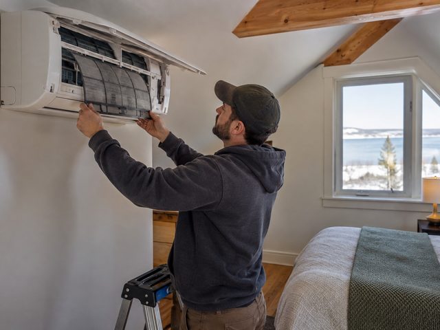 Technician servicing an indoor heat pump head in an upstairs bedroom under a sloped ceiling
